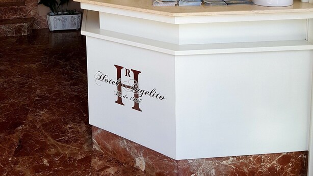 A hotel reception desk with a marble floor, potted plants, and a sign indicating the hotel name and year of establishment. - Hotel Angelito (O Grove)