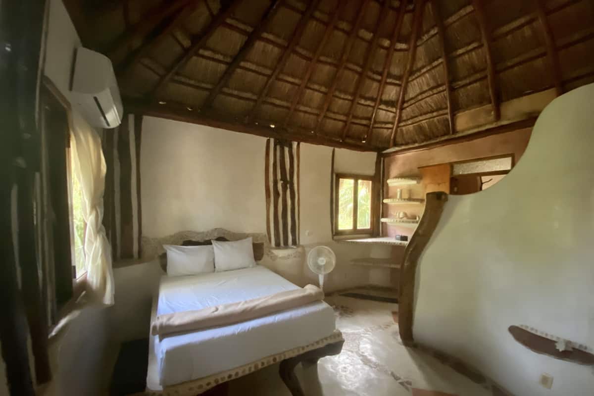 Una habitación con cama, ventilador, ventana y techo de paja.