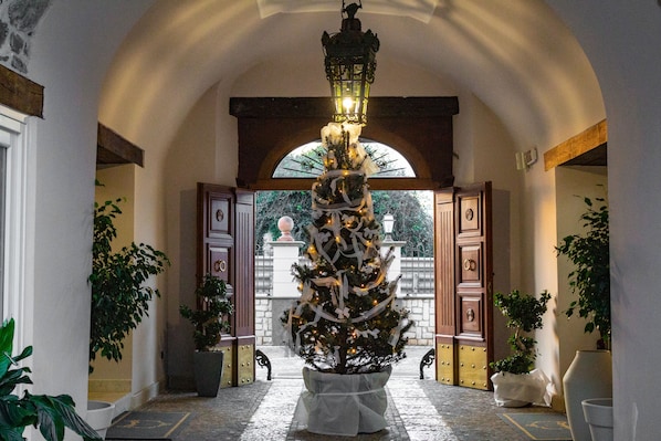 Un amplio pasillo con un gran árbol de Navidad decorado en el centro, flanqueado por plantas en macetas y un arco de piedra.