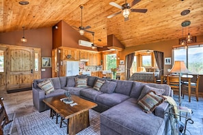 A spacious living area with a large sectional sofa, wooden ceiling, and a view of the mountains through the large windows. - 28 Mi to Yosemite: Lovely Mtn Cottage w/ Deck! (North Fork)