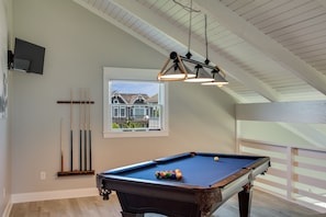 Une table de billard dans une pièce dont le plafond est en pente, une fenêtre donnant sur une maison et un téléviseur fixé au mur.