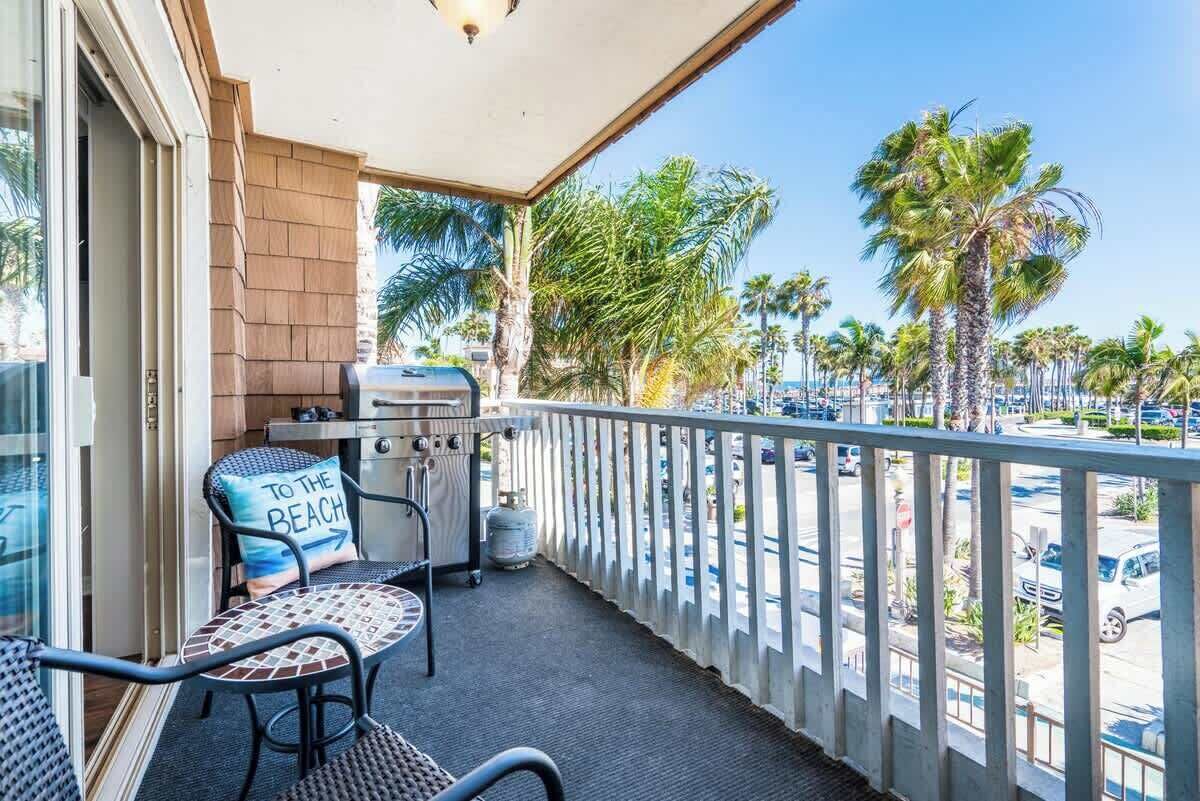 A balcony with a grill, a chair with a pillow, and a view of palm trees and a parking lot.