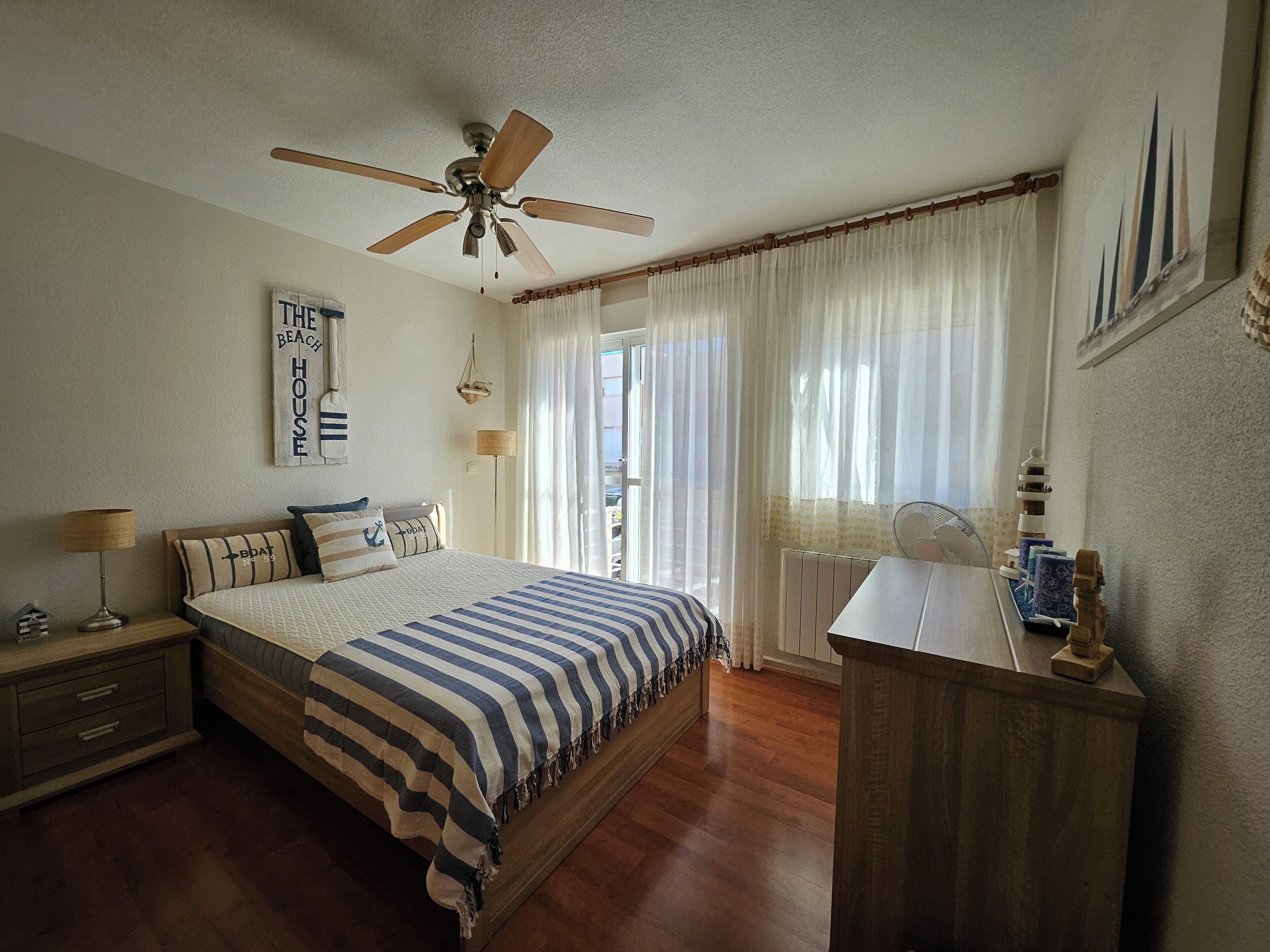 A bedroom with a bed, a nightstand, a desk, a ceiling fan, a window with curtains, and a wall-mounted lamp.
