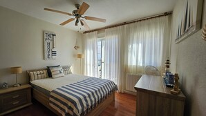 Une chambre à coucher comprenant un lit, une table de chevet, un bureau, un ventilateur de plafond, une fenêtre avec des rideaux et une lampe fixée au mur.
