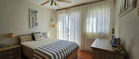 A bedroom with a bed, a nightstand, a desk, a ceiling fan, a window with curtains, and a wall-mounted lamp.