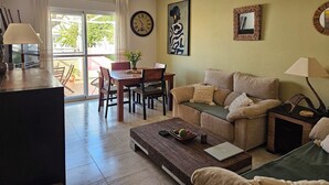 Un salon comprenant un canapé, une table basse, une table à manger et des chaises. Il y a une grande horloge au mur et une fenêtre avec des rideaux.