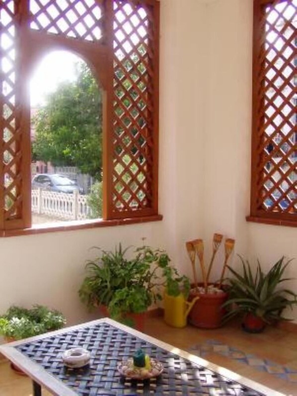 Una habitación con ventanal de celosía, una mesa con mantel tejido y varias plantas en macetas.