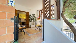 Una habitación con piso de baldosas, puerta de madera, letrero con un número, una planta, una mesa con sillas y una ventana que ofrece vistas al exterior.