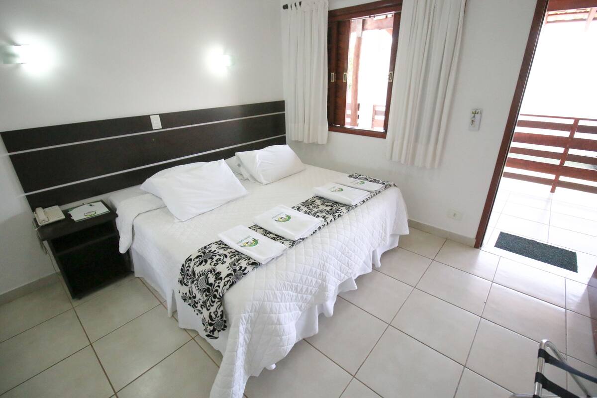 A neatly made bed with white bedding and a patterned runner, a bedside table with a phone and papers, and a window with curtains.