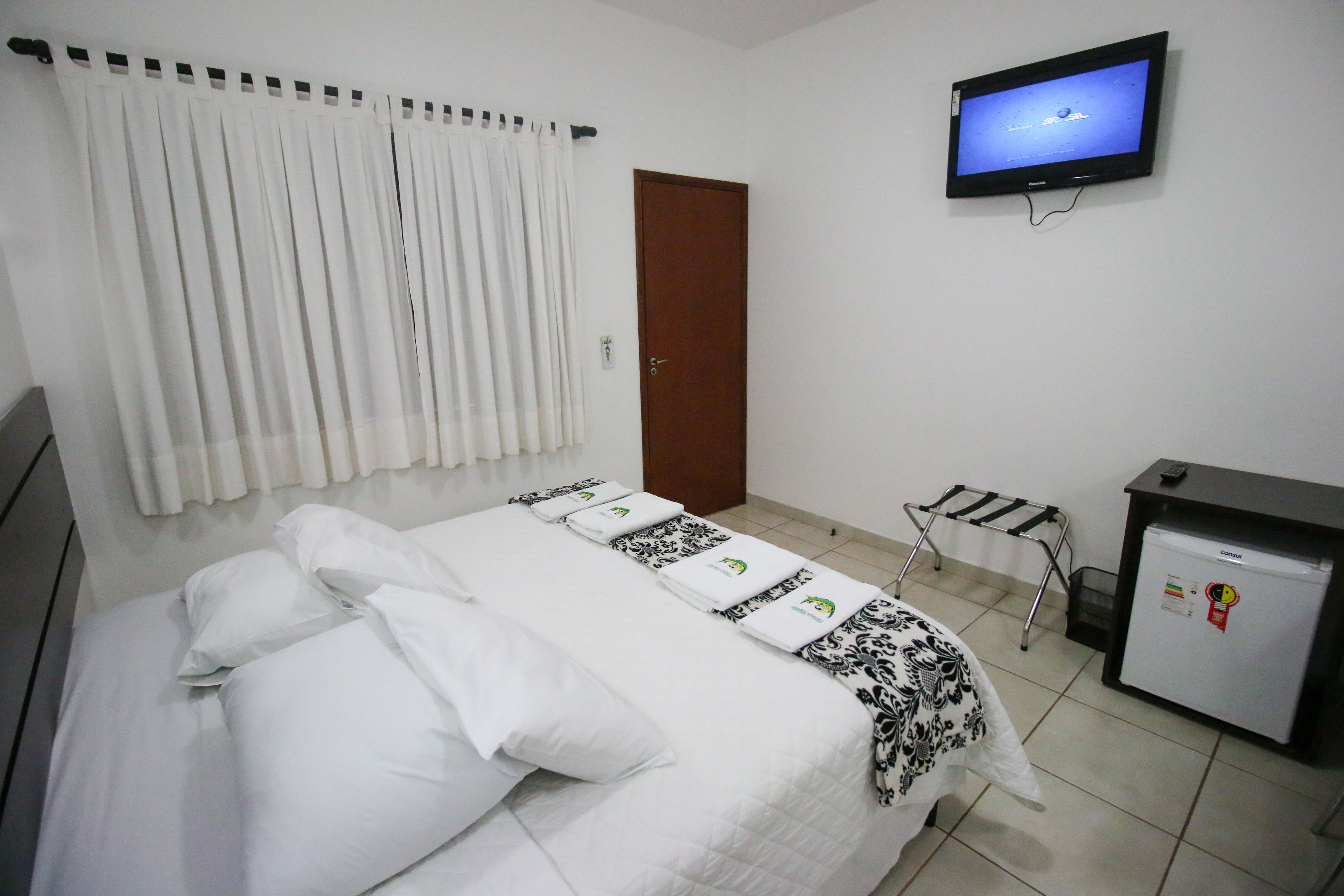 A hotel room with a bed, a television on the wall, a desk with a refrigerator, and a window with curtains.