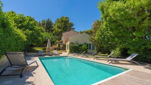 Una piscina rectangular con cubierta de madera, rodeada de exuberante vegetación y una casa al fondo.