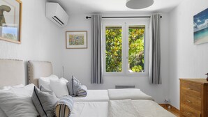 Un dormitorio con dos camas, un aparador de madera, aire acondicionado, una ventana con cortinas e imagen enmarcada en la pared.