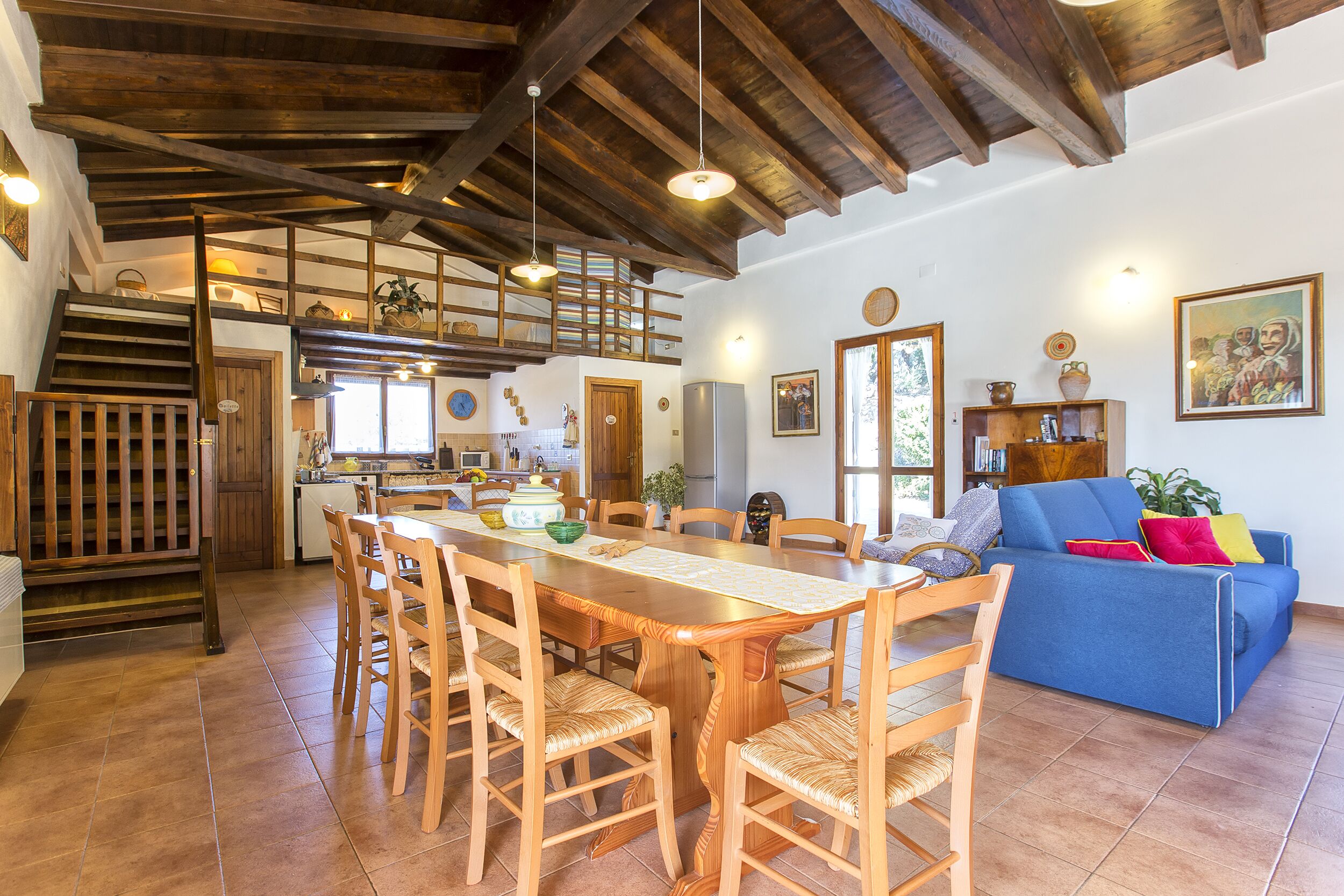 A spacious living area with a wooden dining table and chairs, a blue sofa, and a kitchen in the background.