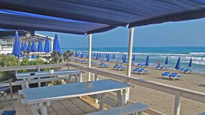 Un patio frente a la playa con mesas de picnic blancas y sombrillas azules, con vista al océano.