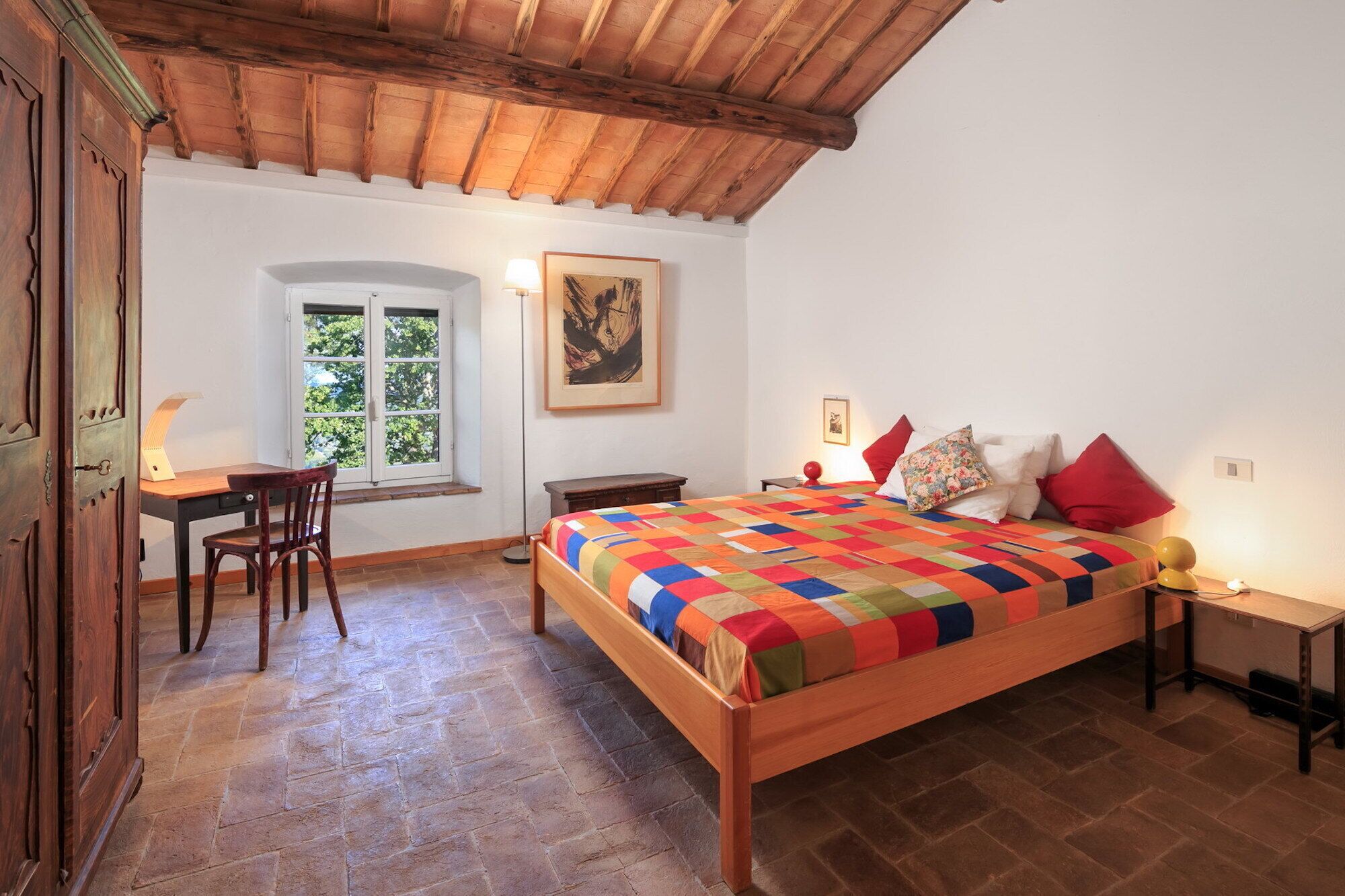 A bedroom with a wooden bed, a desk, and a chair. There is a window with a view of trees.