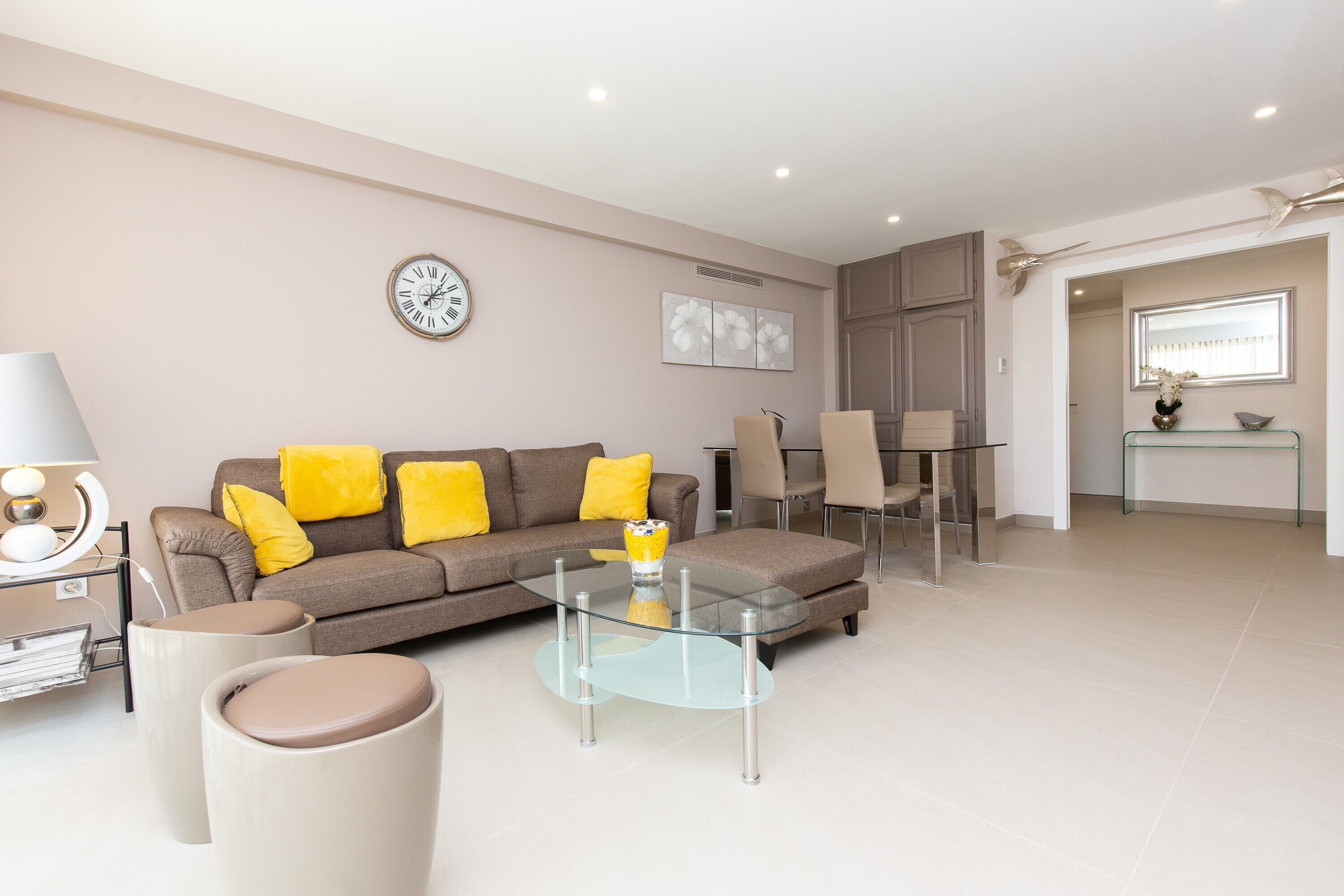 A modern living room with a glass coffee table, a sofa with yellow cushions, a clock on the wall, and a dining area in the background.