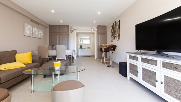 A modern living room with a glass coffee table, a sofa with yellow cushions, a TV on a cabinet, and a dining area in the background.
