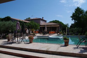 An outdoor pool area with lounge chairs and tables, surrounded by potted plants and a building with a tiled roof.
