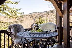 A wooden deck with a round table and two chairs, a bottle of wine, a glass, and a plate with grapes and cheese.