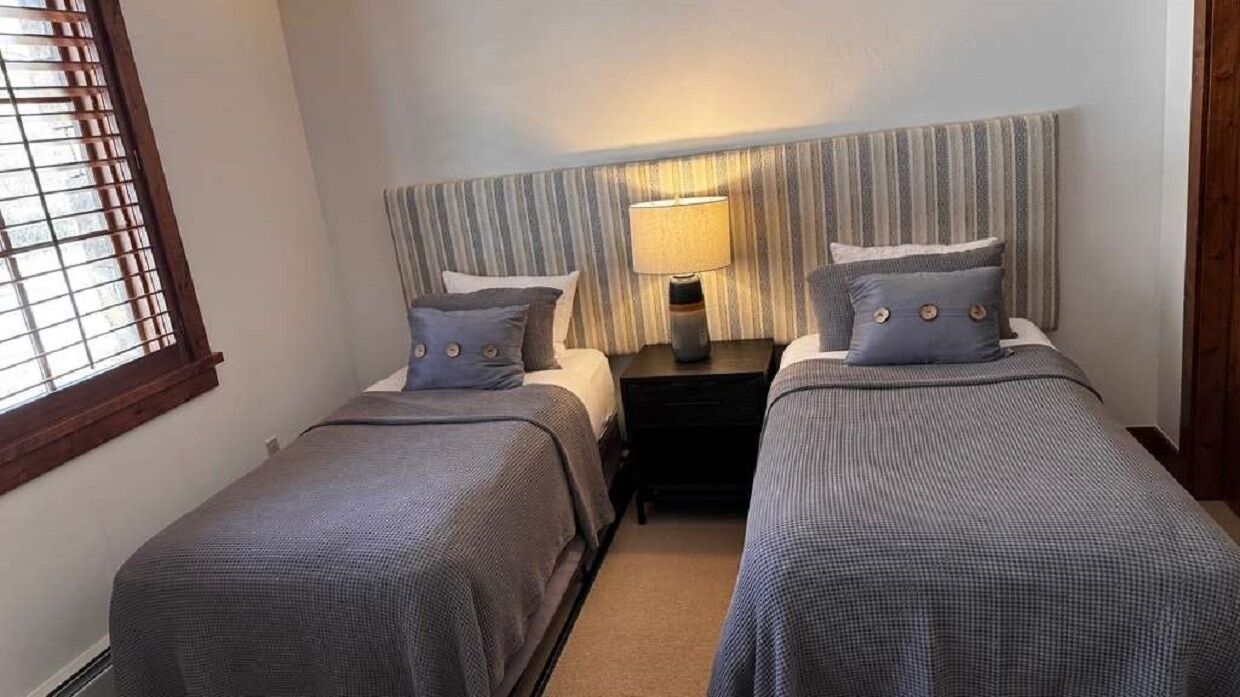 Ein Doppelzimmer mit zwei Betten, einem Nachttisch, einer Lampe und einem Fenster mit Jalousien.