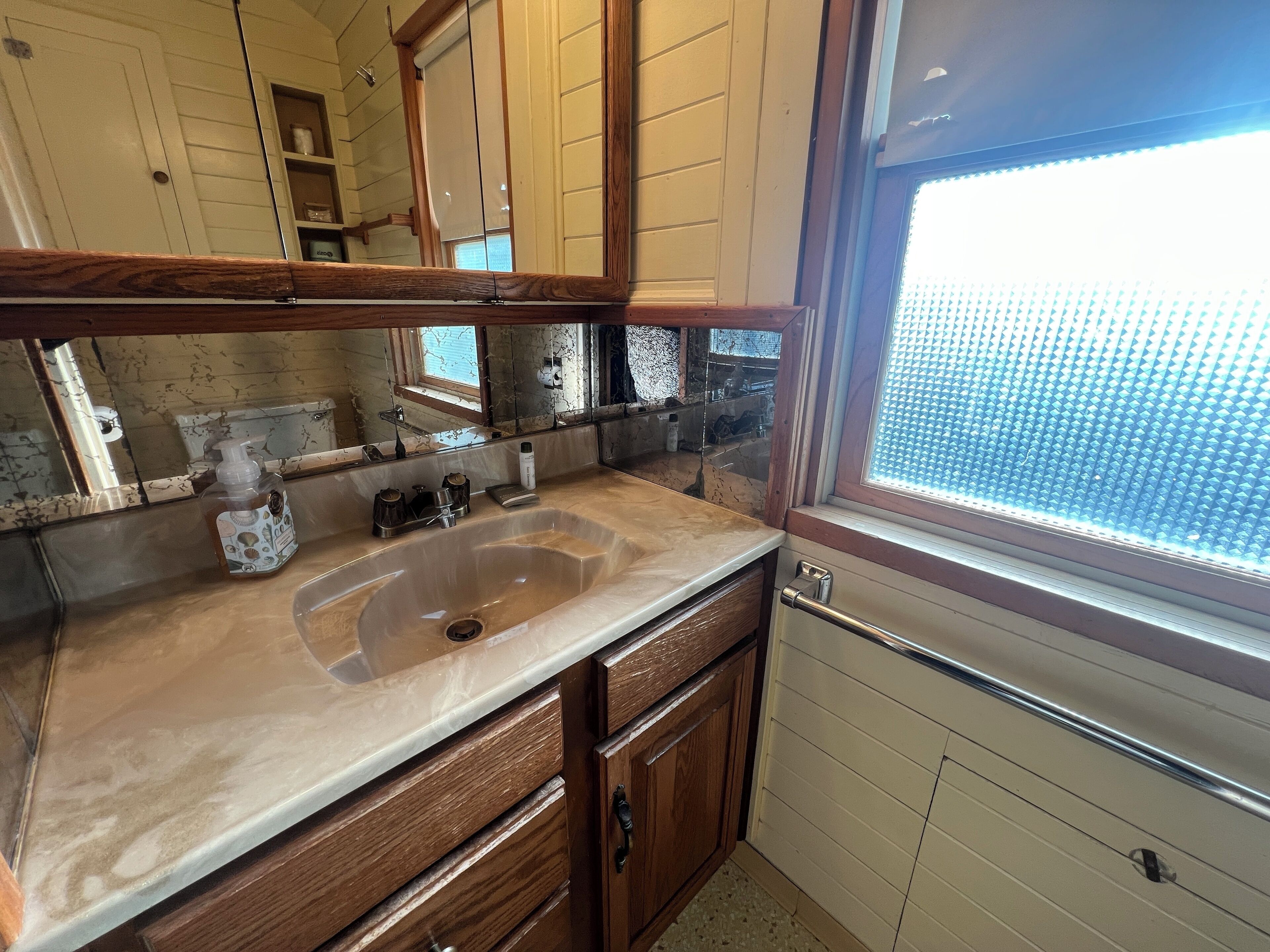 Une salle de bain avec un grand miroir, des meubles en bois, un lavabo et une fenêtre avec des stores.