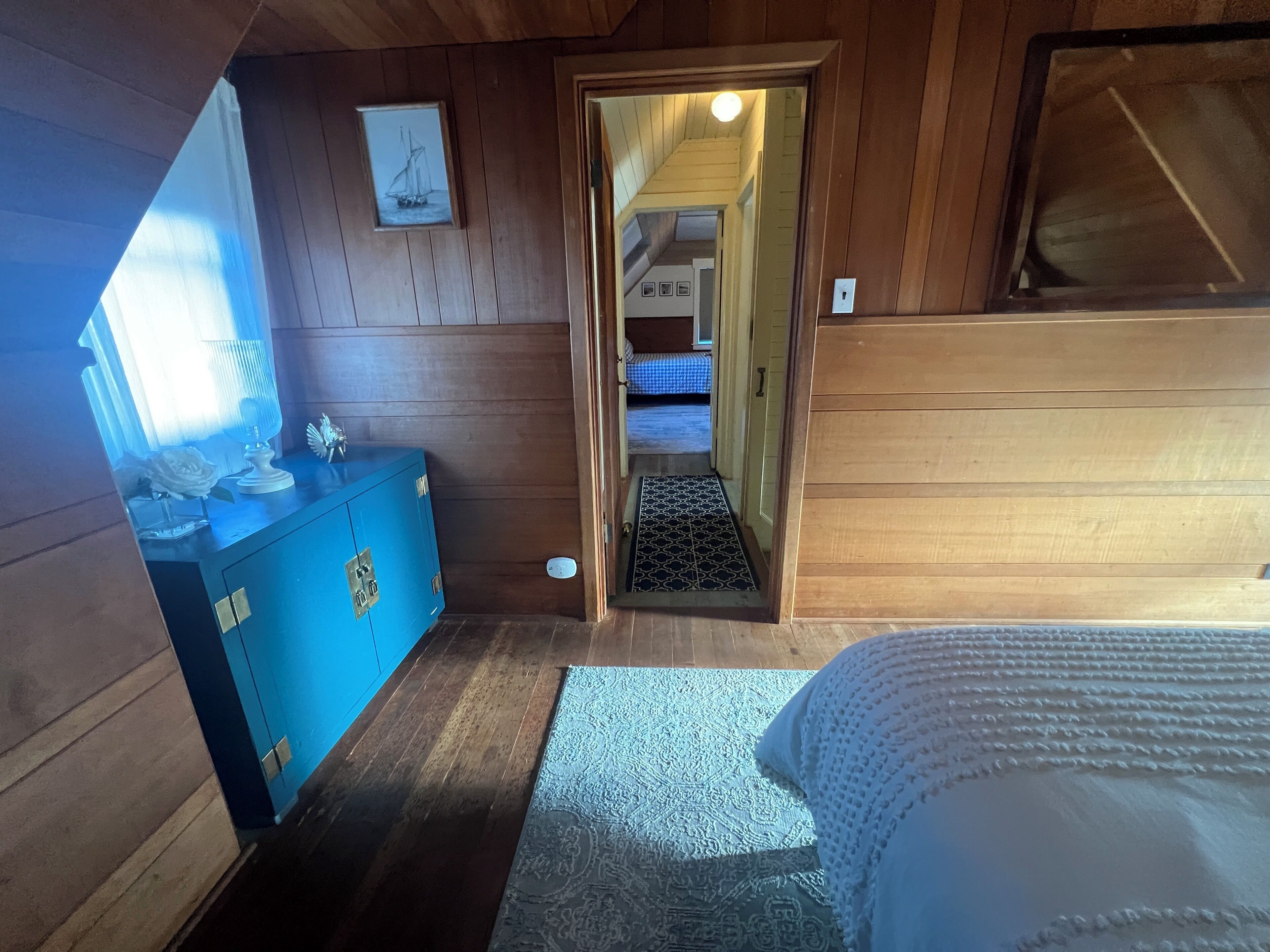 Ein Zimmer mit Holzwänden, einem Bett, einer Kommode, einem gerahmten Bild und einem zur nächsten Kammer führenden Durchgang.