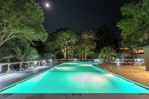 A nighttime swimming pool with clear water, surrounded by trees and lounge chairs.