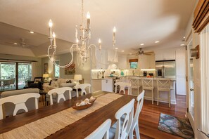 A spacious dining area with a wooden table and white chairs, a chandelier, and a view of the kitchen and living room.