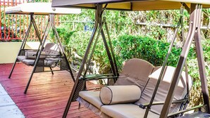 A deck with two swing seats and a cushioned bench, surrounded by greenery. - Brownstone Sea Beach (Nassau)