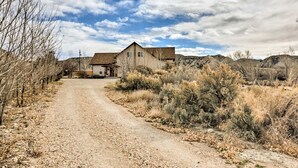A rural road leading to a single-story house with a sloped roof, surrounded by dry vegetation and hills.