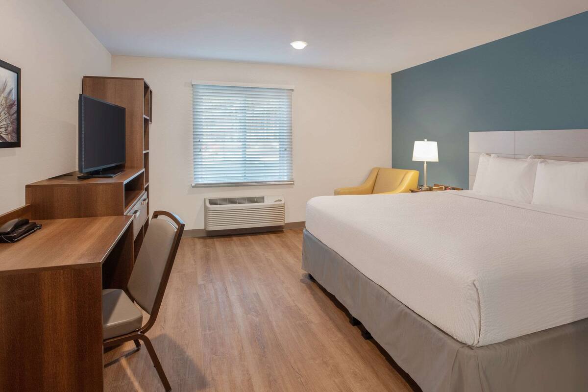 A hotel room with a bed, a desk with a television, a chair, a window with blinds, and a lamp.