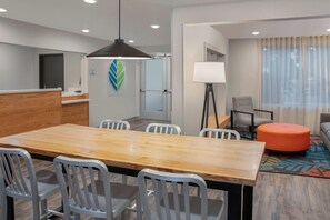 A hotel room with a wooden dining table, white chairs, a black pendant light, a sofa, a floor lamp, and a window with curtains. - WoodSpring Suites Washington DC East Arena Drive (Hyattsville)
