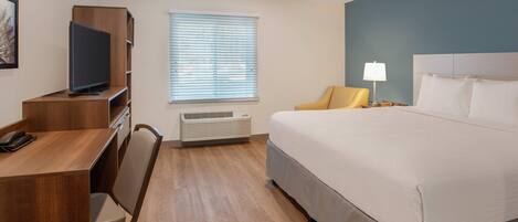 A hotel room with a bed, a desk with a television, a chair, a window with blinds, a bedside lamp, and an air conditioning unit.