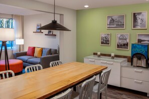 A modern room with a wooden dining table, white chairs, a sofa with colorful cushions, a hanging pendant light, and a wall with framed pictures. - WoodSpring Suites Washington DC East Arena Drive (Hyattsville)