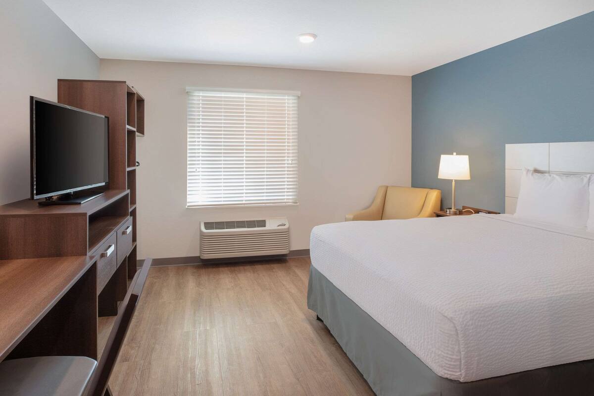 A hotel room with a bed, a TV, a desk, a chair, and a window with blinds.