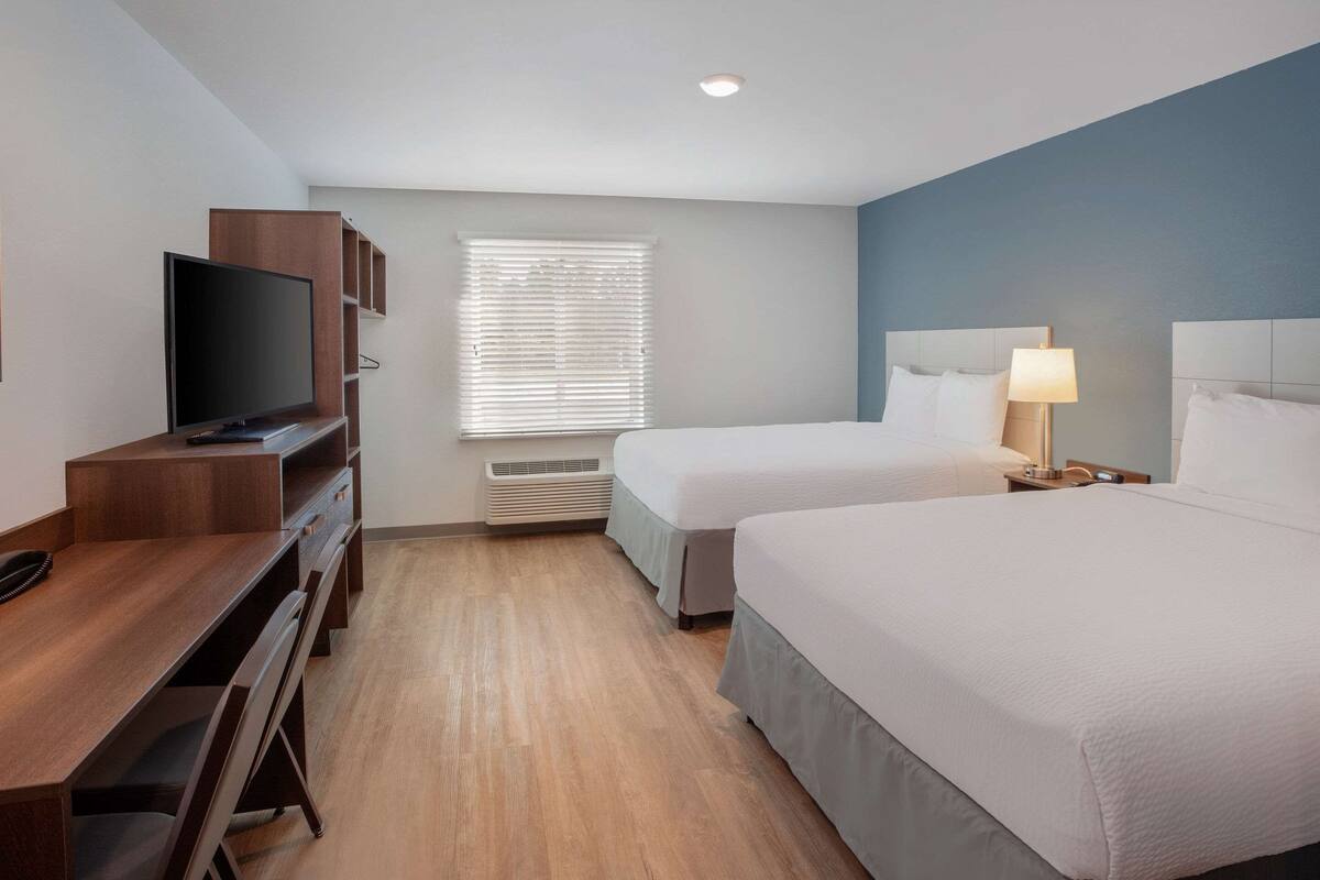 A hotel room with a wooden desk, a television, two beds, a window with blinds, and a nightstand with a lamp.