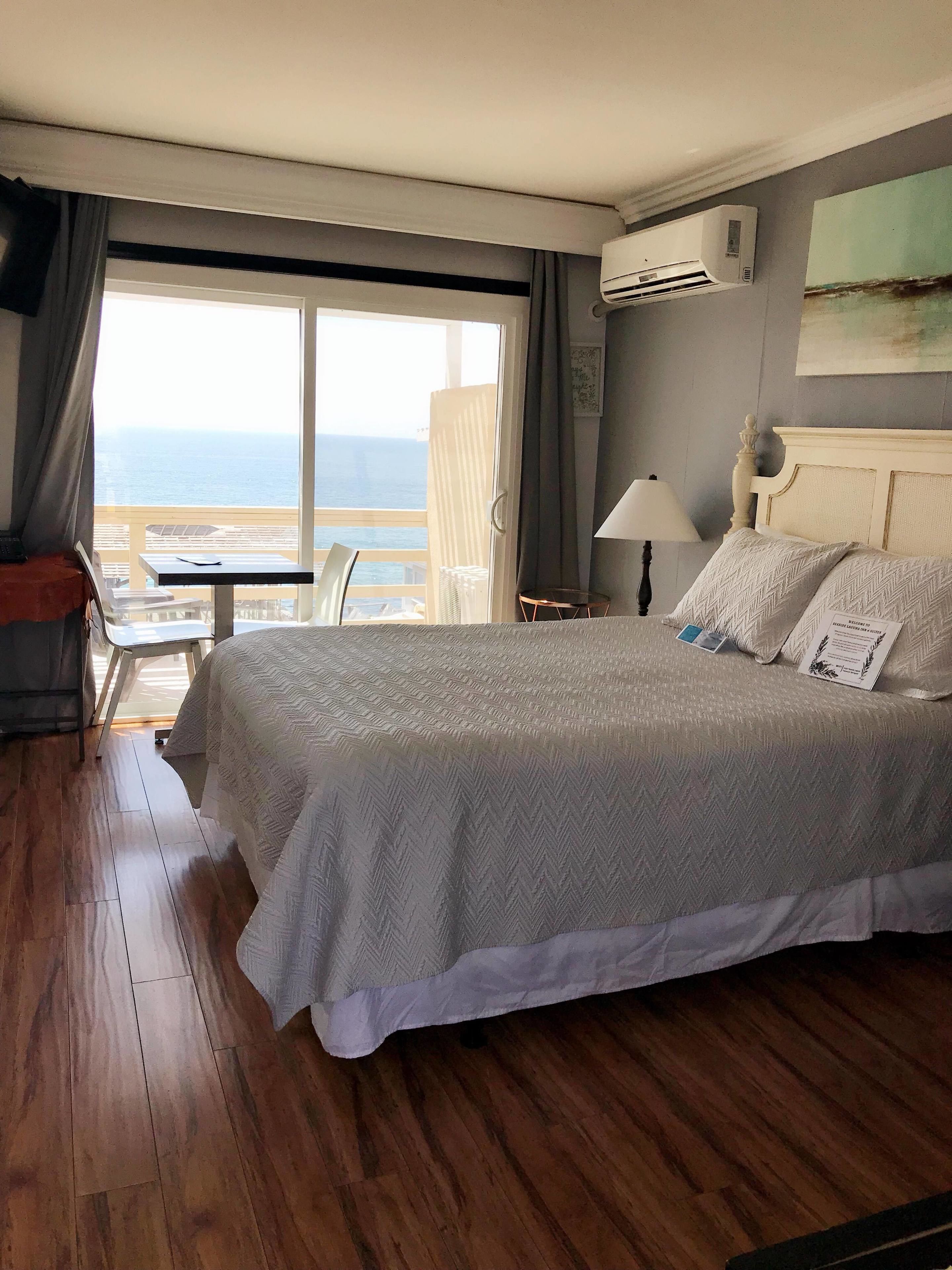 Quarto de hotel com uma cama grande, uma mesa pequena, uma cadeira e vista para o mar através de uma janela ampla.