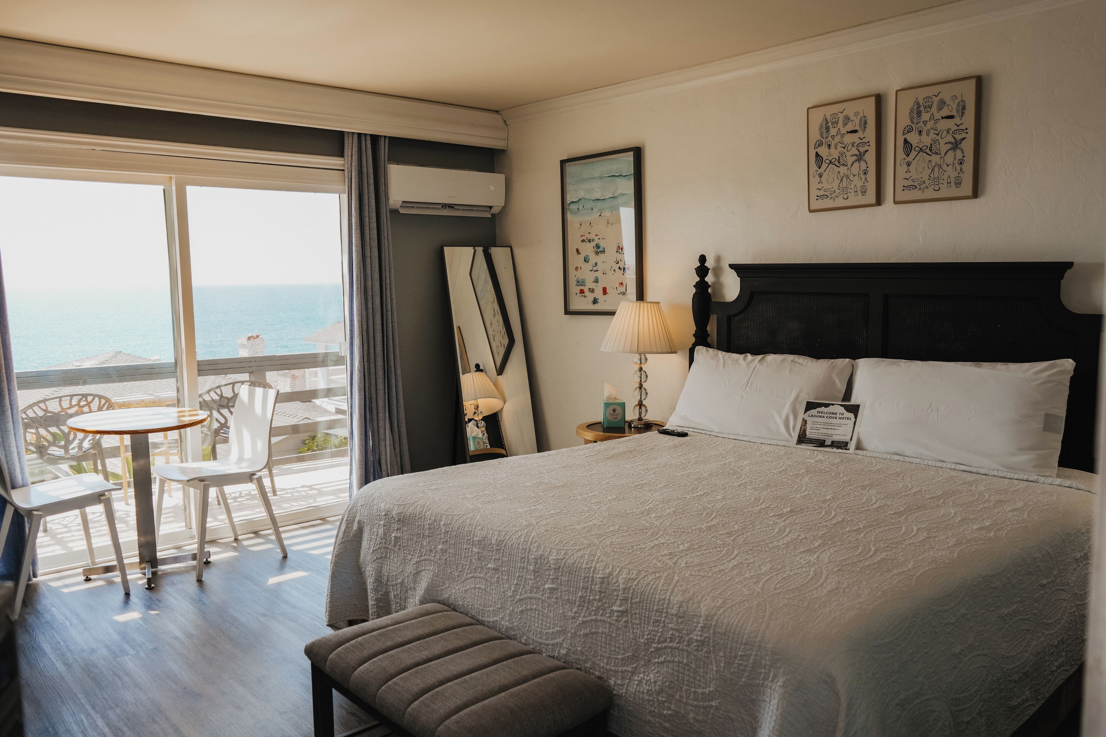 A hotel room with a large bed, a bedside table with a lamp, a small round table with chairs, and a view of the sea through the window.