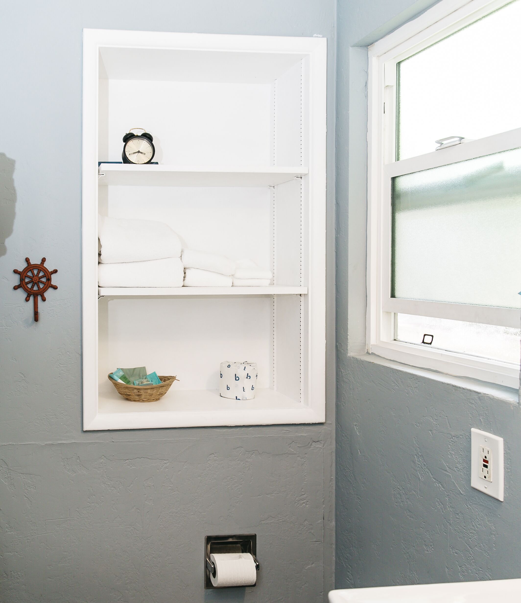 Un bagno con un mobiletto contenitore bianco, una decorazione a forma di timone da nave, un cesto e un rotolo di carta igienica.