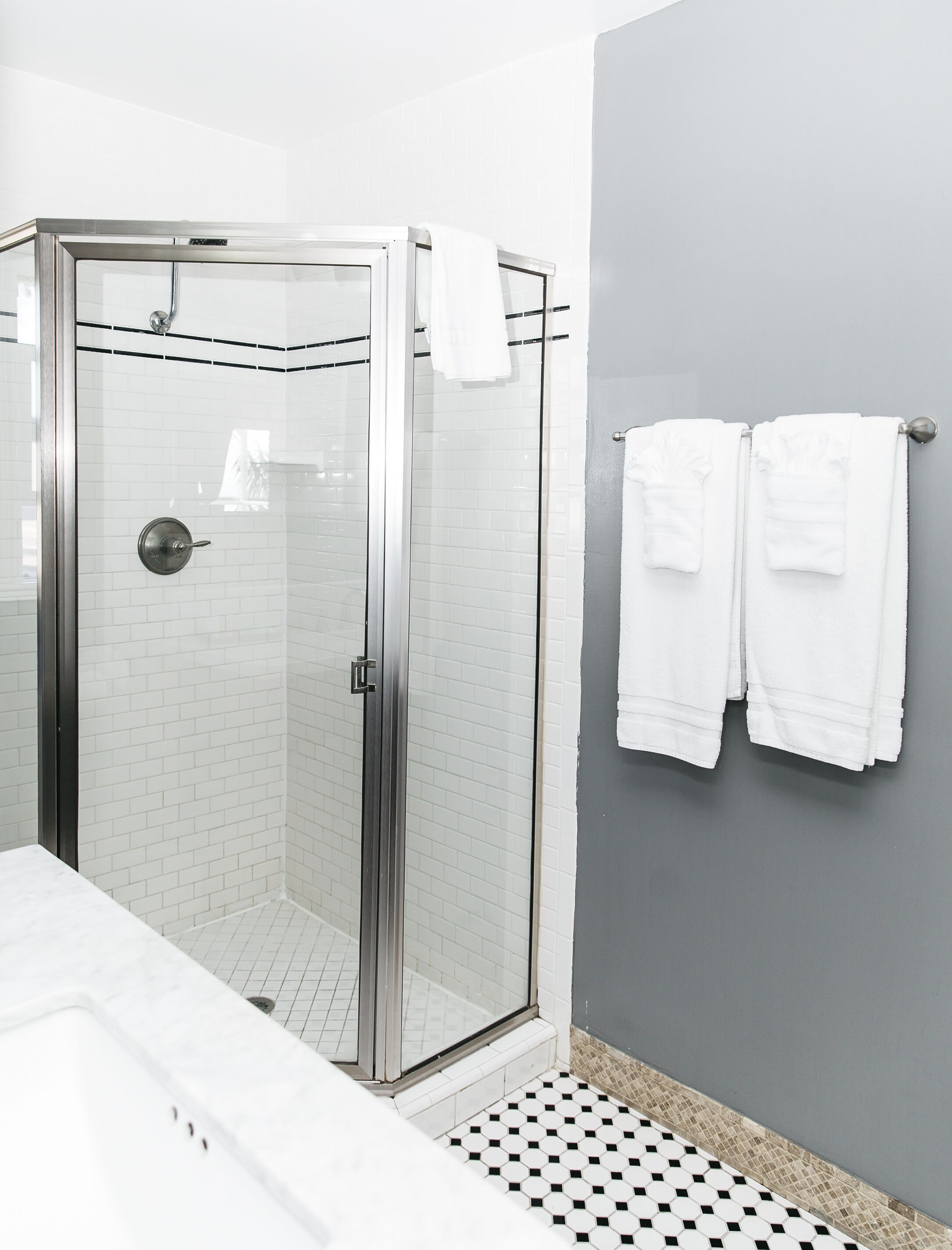 A shower with a glass door, white tiled walls, and a gray accent wall. There are two white towels hanging on a towel rack.