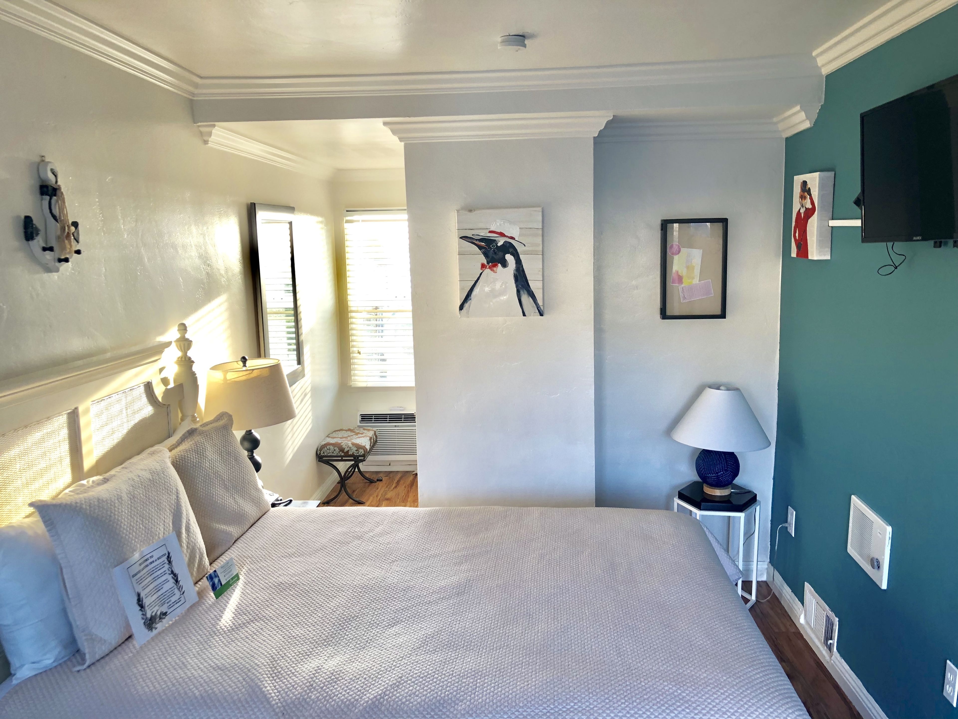 A bedroom with a large bed, a bedside table with a lamp, a wall-mounted TV, and a window with blinds.