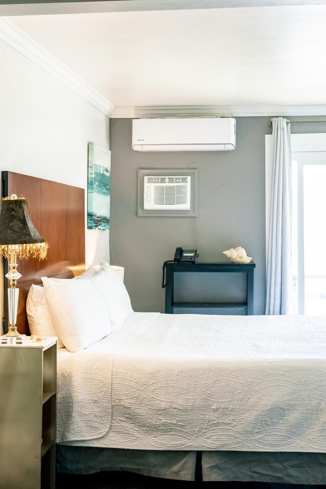 Ein Hotelzimmer mit Bett, Nachttischlampe, Klimaanlage, Nachttisch mit Telefon und einer Dekoration sowie einem Fenster mit Vorhängen.
