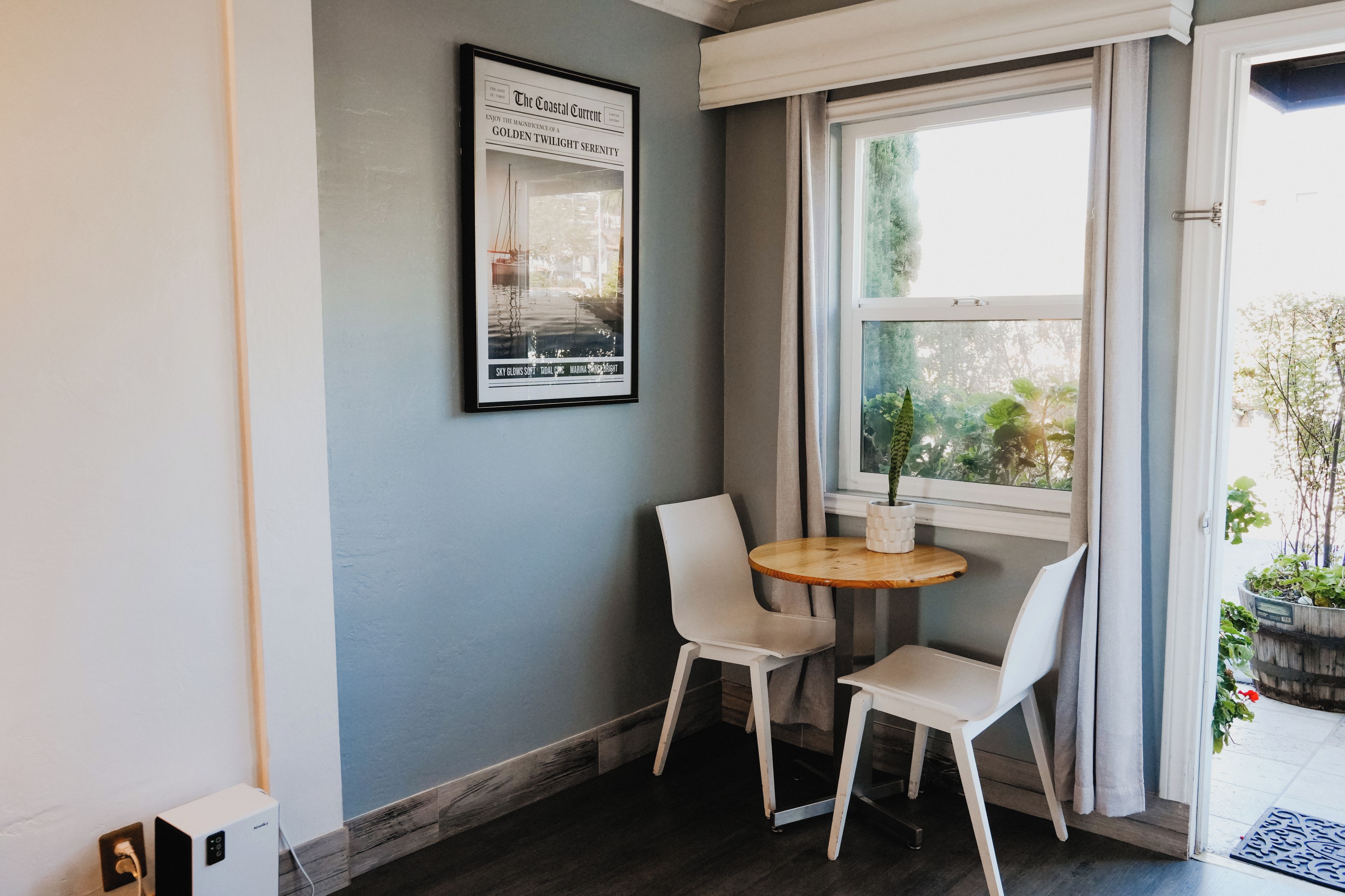 Ein Eckzimmer mit Fenster, zwei Stühlen, einem kleinen Tisch und einem gerahmten Bild an der Wand.