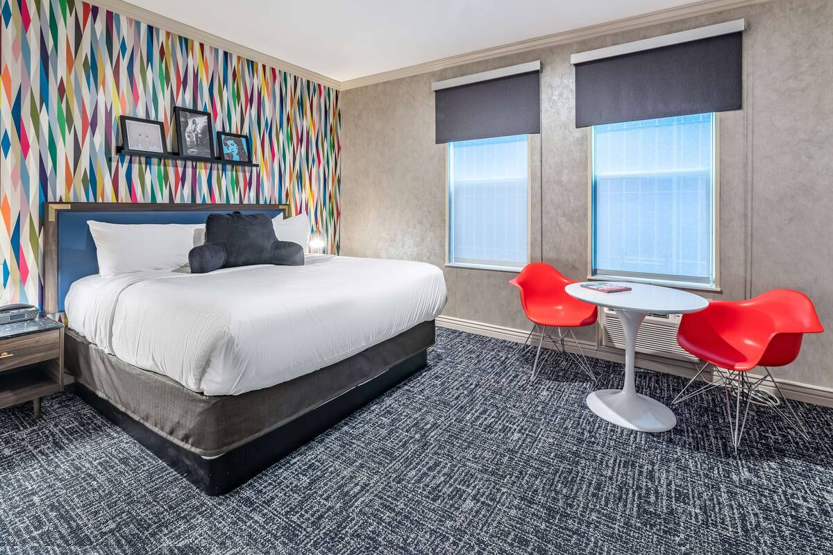 A hotel room with a large bed, a colorful wall, a window with blinds, a small table with red chairs, and a carpeted floor.