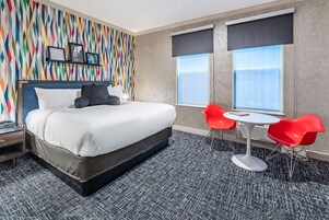 Una habitación de hotel con una cama grande, una pared colorida, una ventana con persianas, una pequeña mesa con sillas rojas y suelo alfombrado.