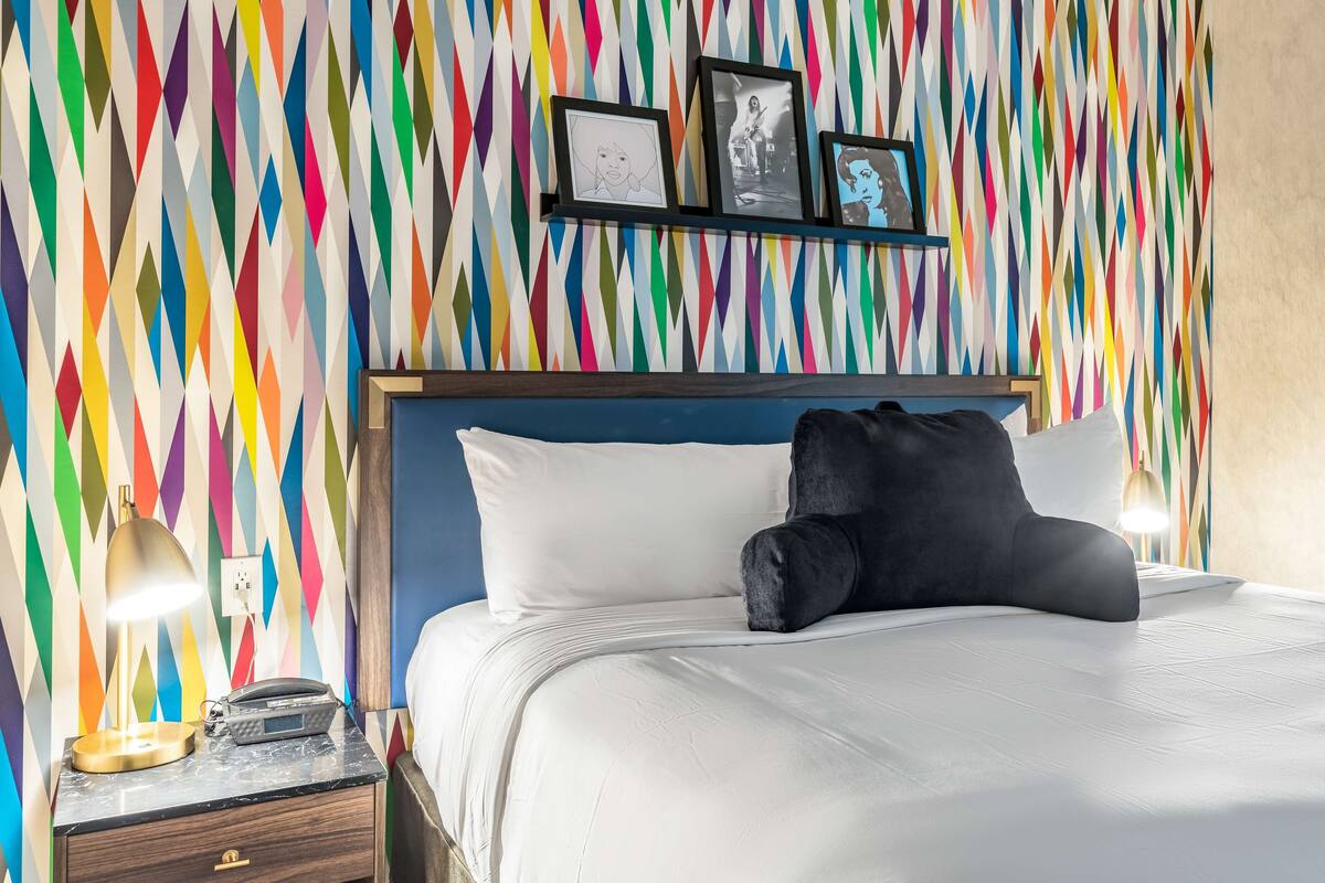 A hotel room with a colorful geometric-patterned wall, a bed with white linens, a black pillow, a bedside lamp, and a nightstand with a phone and a lamp.