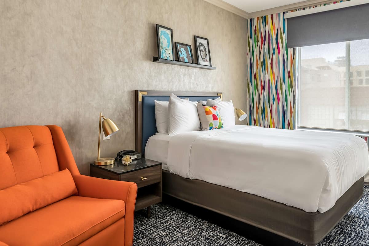 A hotel room with a large bed, a nightstand, a lamp, a chair, and a window with a view of buildings.