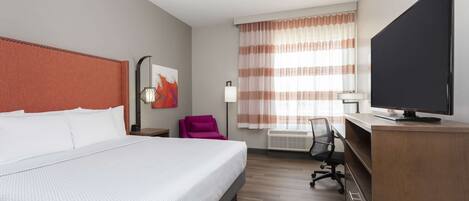 Ein Hotelzimmer mit einem großen Bett, einem Schreibtisch mit Computer, einem Sessel, einem Fernseher und einem Fenster mit Vorhängen.