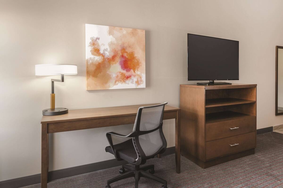 A hotel room with a wooden desk, a black office chair, a flat-screen TV, a wall-mounted lamp, and a large abstract painting.