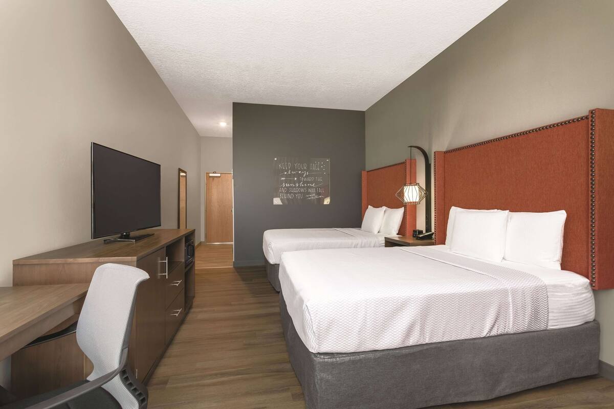 A hotel room with two beds, a desk with a TV, a chair, and a motivational quote on the wall.